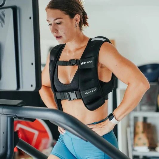 Female athlete working out on the STEPR Pro+ Stair Climber in a garage, with a functional trainer in the background and wearing a weighted vest.