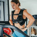 Female athlete working out on the STEPR Pro+ Stair Climber in a garage, with a functional trainer in the background and wearing a weighted vest.