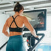 Close-up from the backside of a female athlete exercising on the STEPR Pro+ Stair Climber, gripping the handle with the large 27” HD touchscreen prominently displayed.